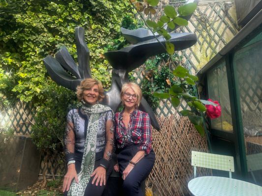 Claudia Turbay, Director of External Affairs at SSCC, and Hedva Ser, UNESCO Goodwill Ambassador for Cultural Diplomacy, seated on a bench next to the large Tree of Peace sculpture, a metal tree-shaped artwork symbolizing peace, in an outdoor setting.